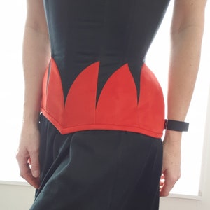 May include: A black corset with a red, pointed, scalloped bottom. The corset is made of a smooth fabric and has a fitted silhouette.