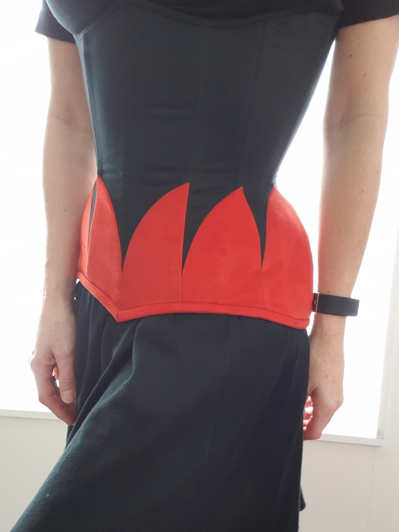 May include: A black corset with a red, pointed, scalloped bottom. The corset is made of a smooth fabric and has a fitted silhouette.