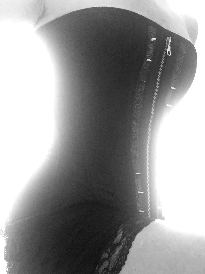 Puede incluir: Un cors&eacute; negro sin tirantes con una cremallera en la parte delantera y tachuelas plateadas a lo largo de la cremallera. El cors&eacute; tiene un ribete de encaje en la parte inferior.