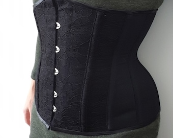 Op de afbeelding: Een zwart kant-corset met zilveren metalen oogjes en een satijnen afwerking. Het corset wordt gedragen over een groen shirt.