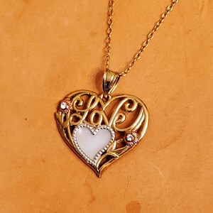 Op de afbeelding: Gouden ketting met een hartvormige hanger met het woord "love" in een decoratief schrift. De hanger heeft een wit parelmoer centrum en kleine heldere stenen.
