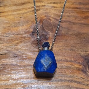 May include: A faceted, dark blue lapis lazuli pendant necklace. The gemstone bottle-shaped pendant hangs from a silver-colored chain. The pendant has a small, matching blue stopper. The background is a wooden surface.