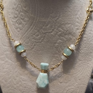 May include: A gold-toned necklace featuring a small, light blue perfume bottle pendant. The necklace has a delicate chain with light blue and white beads. The pendant is a light blue, hexagonal shape with a gold-colored band around the top.