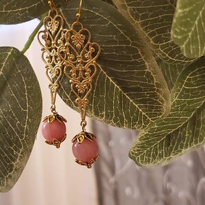 May include: Gold-colored filigree earrings with pink, round beads. The earrings have an ornate, openwork design with heart and floral motifs. The pink beads are suspended from the filigree. The earrings are hanging in front of green leaves.