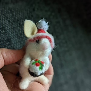 Peut inclure: Souris en feutre blanc portant un bonnet de Père Noël rouge et blanc avec un pompon blanc, tenant un pudding de Noël en feutre blanc avec du houx vert et des baies rouges.
