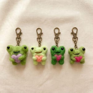 May include: Four handmade frog keyrings in shades of green, each holding a heart. The keyrings have black eyes and are attached to bronze-coloured clasps. The frogs are made of a fuzzy material.