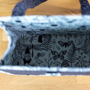 May include: Open view of a tote bag with a dark gray exterior and a patterned interior. The interior fabric features black and white illustrations of butterflies, dragonflies, gnomes, and foliage. The bag has a handle.