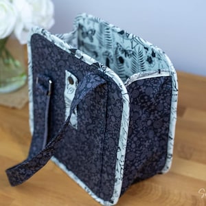 May include: A navy blue tote bag with a floral pattern. The bag has a light blue interior with a botanical print. The bag has a handle and a silver-colored metal clasp. The bag is sitting on a wooden surface.