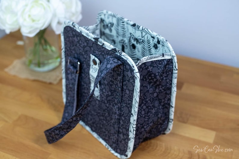May include: A navy blue tote bag with a floral pattern. The bag has a light blue interior with a botanical print. The bag has a handle and a silver-colored metal clasp. The bag is sitting on a wooden surface.