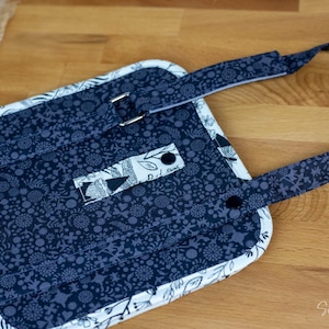May include: A navy blue and white quilted apron with a floral pattern. The apron has a strap with a metal buckle and a smaller strap with two black buttons. The edges are trimmed with white fabric.