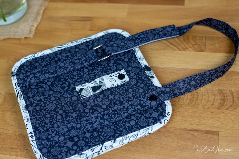 May include: A navy blue and white quilted apron with a floral pattern. The apron has a strap with a metal buckle and a smaller strap with two black buttons. The edges are trimmed with white fabric.
