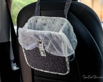 May include: A car trash bag made of black and white floral patterned fabric, hanging from a car seat. The bag has a clear plastic liner and a fabric strap for hanging. The bag is designed to hold trash and keep the car clean.