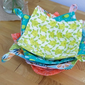 Könnte beinhalten: Ein Stapel bunter Stoff-Schalenuntersetzer mit Schlaufengriffen. Die Untersetzer weisen verschiedene Muster auf, darunter Schildkröten, Ananas und florale Designs. Die Farben umfassen Türkis, Orange, Rosa und Grün.