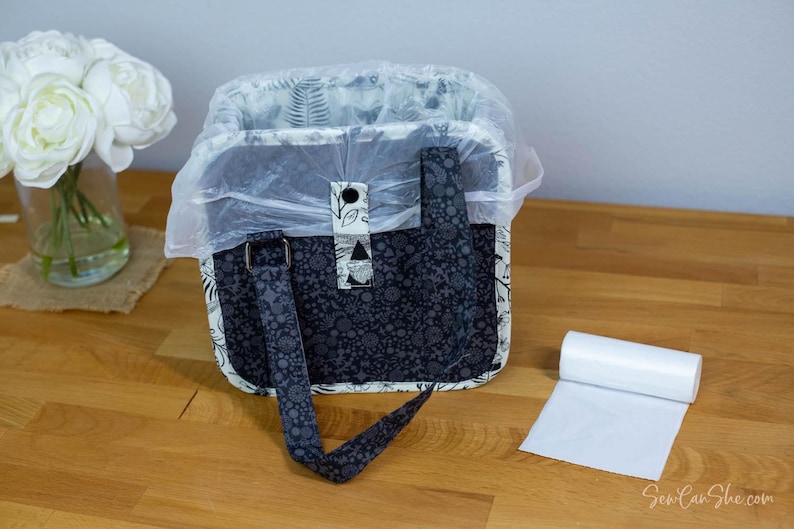May include: A patterned tote bag with a plastic liner, featuring a black and gray floral design. The bag has a handle and a buckle. A roll of white plastic bags sits nearby. The bag is on a wooden surface.