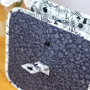 May include: A handmade, square-shaped bag with a dark gray floral pattern on the front and a black button closure. The interior and trim feature a contrasting black and white botanical print. The bag is open, revealing the interior.