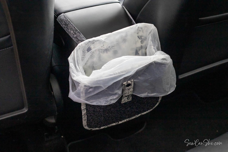 May include: A car trash bag with a patterned exterior and a clear plastic liner. The bag is designed to hang from the back of a car seat. The bag has a decorative clasp.