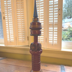 May include: A tall, decorative wooden object with a dark blue pointed top, stacked brown rings, and a red brick-like base. The base has a dark green and tan color. The object is displayed on a wooden surface near a window with white shutters.