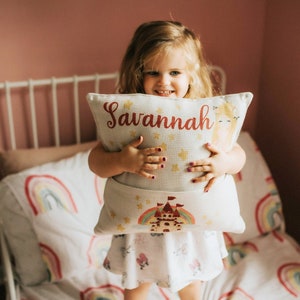 May include: A white pillow with a rainbow and castle design. The pillow has a pocket on the back and the name "Savannah" embroidered on the front in red.