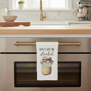 May include: A white waffle-weave kitchen towel hanging on an oven handle. The towel features the black text "DON'T GET ME started" above a graphic illustration of a glass jar filled with sourdough starter covered by a cloth lid.