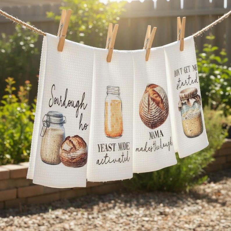 May include: Four white waffle-weave kitchen towels hang on a clothesline, each clipped with a wooden clothespin. The towels feature watercolor illustrations of sourdough starters and bread, with text that reads "Sourdough ho", "Yeast mode activated", "Mama makes the dough", and "Don't get me started".