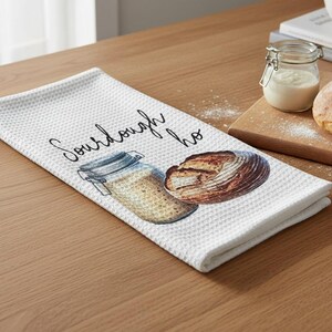 May include: White waffle-weave kitchen towel with a watercolor illustration of a sourdough loaf and a jar of starter. The towel has the word "Sourdough" printed on it in a script font. A loaf of bread and a jar of starter are on a wooden cutting board.