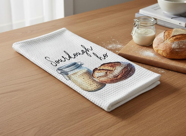 May include: White waffle-weave kitchen towel with a watercolor illustration of a sourdough loaf and a jar of starter. The towel has the word "Sourdough" printed on it in a script font. A loaf of bread and a jar of starter are on a wooden cutting board.