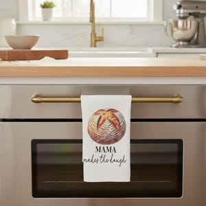 May include: White kitchen towel with a watercolor image of a loaf of bread. The towel is hanging on a gold-colored oven handle. Text on the towel reads "MAMA makes the dough".