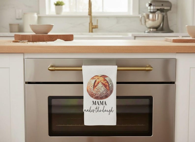 May include: White kitchen towel with a watercolor image of a loaf of bread. The towel is hanging on a gold-colored oven handle. Text on the towel reads "MAMA makes the dough".