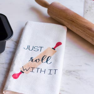 May include: A white kitchen towel with the text "JUST roll WITH IT" and a red and brown rolling pin graphic.