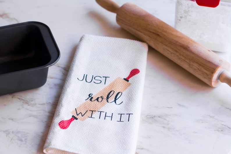 May include: A white kitchen towel with the text "JUST roll WITH IT" and a red and brown rolling pin graphic.