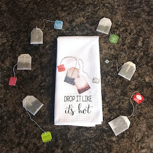 May include: White kitchen towel with a graphic of tea bags and the text "DROP IT LIKE it's hot".  The towel is surrounded by various tea bags in different colors.