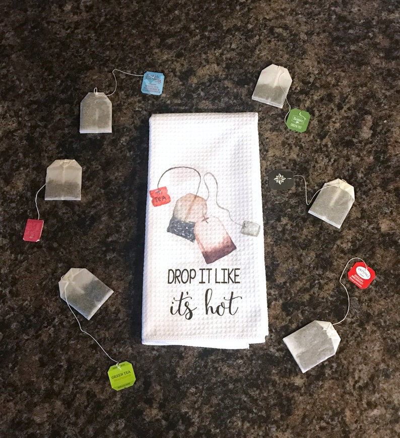 May include: White kitchen towel with a graphic of tea bags and the text "DROP IT LIKE it's hot".  The towel is surrounded by various tea bags in different colors.