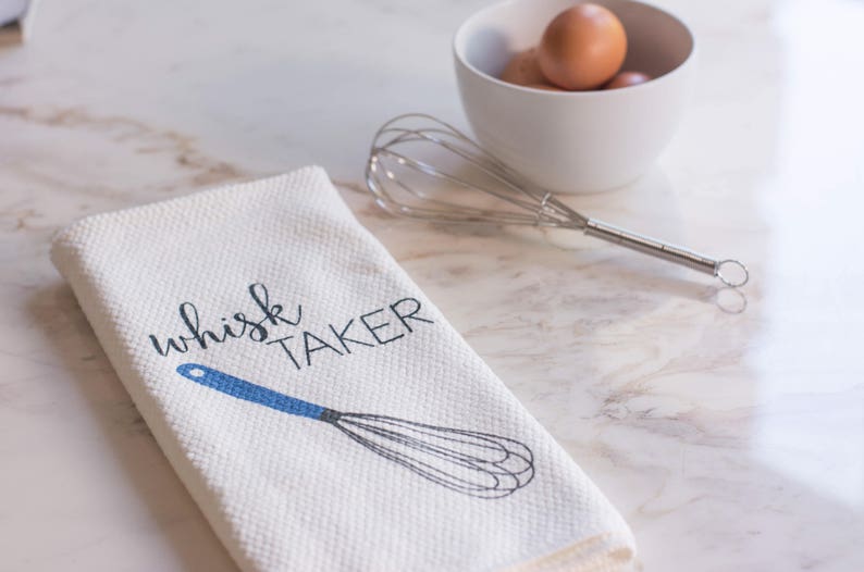 May include: White kitchen towel with blue text that reads "whisk taker" and a blue and gray whisk illustration.