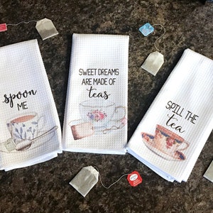 May include: Three white tea towels with teacup designs and sayings. The first towel says "Spoon Me" with a teacup and spoon. The second towel says "Sweet Dreams Are Made Of Teas" with a teacup and a tea strainer. The third towel says "Spill The Tea" with a teacup.