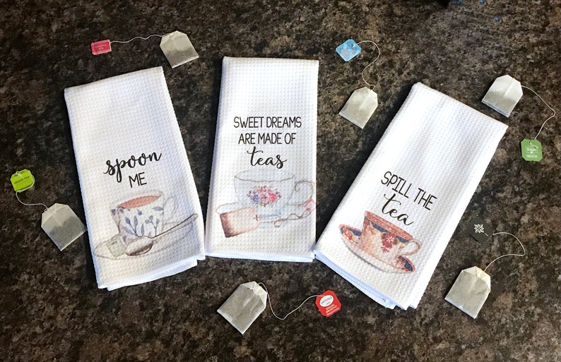 May include: Three white tea towels with teacup designs and sayings. The first towel says "Spoon Me" with a teacup and spoon. The second towel says "Sweet Dreams Are Made Of Teas" with a teacup and a tea strainer. The third towel says "Spill The Tea" with a teacup.
