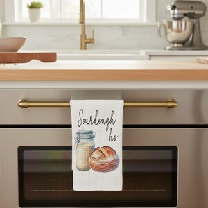 May include: A white waffle-weave kitchen towel hanging from a stainless steel oven handle. The towel features a watercolor illustration of a glass sourdough starter jar and a round loaf of bread with the black cursive text Sourdough ho.
