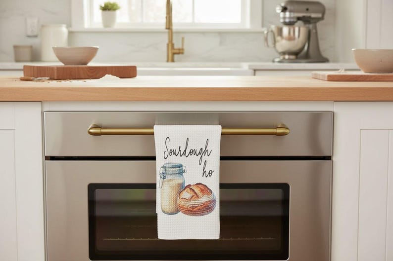 May include: A white waffle-weave kitchen towel hanging from a stainless steel oven handle. The towel features a watercolor illustration of a glass sourdough starter jar and a round loaf of bread with the black cursive text Sourdough ho.