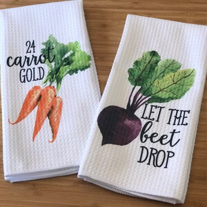 K&ouml;nnte beinhalten: Zwei wei&szlig;e K&uuml;chent&uuml;cher mit Aquarell-Gem&uuml;semotiven und Wortspielen. Das erste Handtuch zeigt zwei orangefarbene Karotten mit gr&uuml;nen Bl&auml;ttern und dem Text "24 Karat Gold". Das zweite Handtuch zeigt eine violette Rote Bete mit gr&uuml;nen Bl&auml;ttern und dem Text "Let the beet drop".