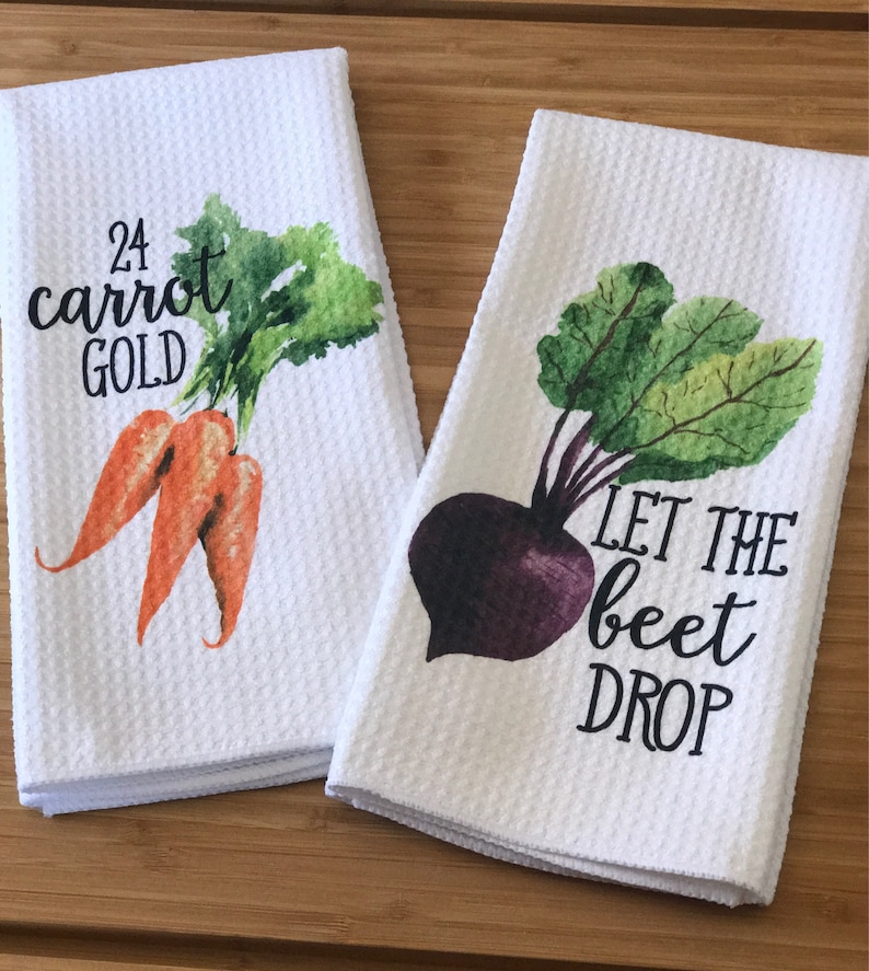 K&ouml;nnte beinhalten: Zwei wei&szlig;e K&uuml;chent&uuml;cher mit Aquarell-Gem&uuml;semotiven und Wortspielen. Das erste Handtuch zeigt zwei orangefarbene Karotten mit gr&uuml;nen Bl&auml;ttern und dem Text "24 Karat Gold". Das zweite Handtuch zeigt eine violette Rote Bete mit gr&uuml;nen Bl&auml;ttern und dem Text "Let the beet drop".