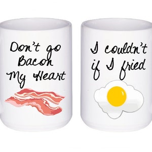 May include: Two white ceramic mugs with black text and illustrations. The mug on the left says "Don't go Bacon my Heart" and has a cartoon image of bacon. The mug on the right says "I couldn't if I fried" and has a cartoon image of a fried egg.