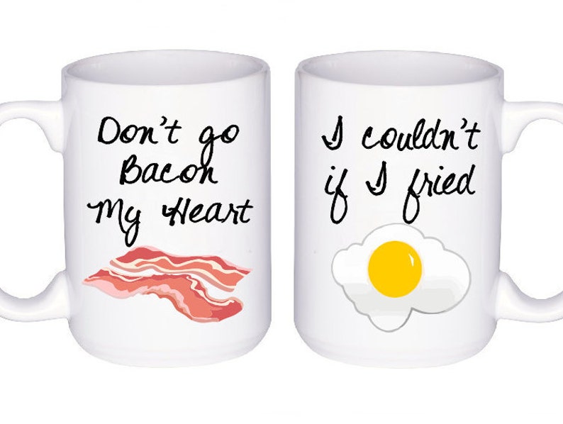 May include: Two white ceramic mugs with black text and illustrations. The mug on the left says "Don't go Bacon my Heart" and has a cartoon image of bacon. The mug on the right says "I couldn't if I fried" and has a cartoon image of a fried egg.
