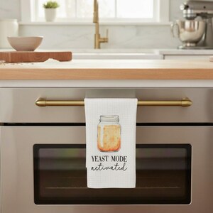 May include: White kitchen towel with a watercolor illustration of a jar with yeast and the text "YEAST MODE activated". The towel is hanging on a gold-colored oven handle. The kitchen has a light wood countertop.