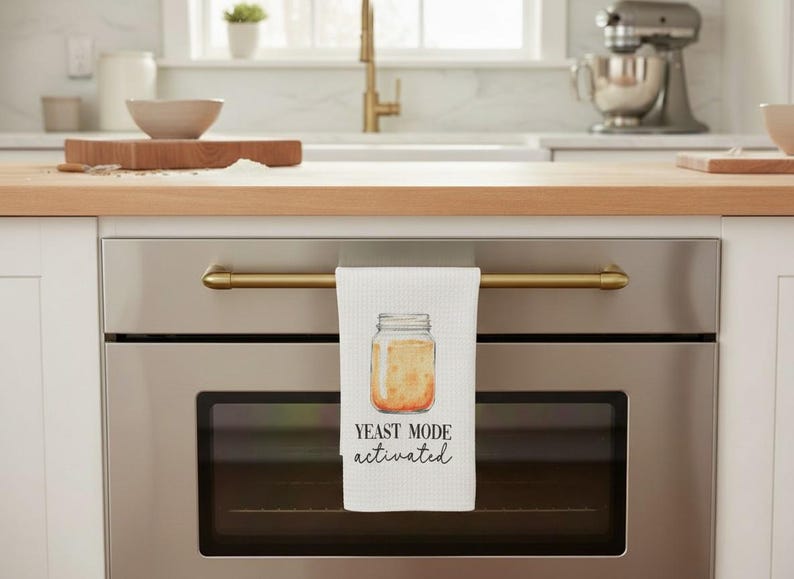 May include: White kitchen towel with a watercolor illustration of a jar with yeast and the text "YEAST MODE activated". The towel is hanging on a gold-colored oven handle. The kitchen has a light wood countertop.