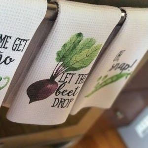 K&ouml;nnte beinhalten: Drei wei&szlig;e K&uuml;chent&uuml;cher mit verschiedenen Gem&uuml;semotiven und Spr&uuml;chen. Das erste Handtuch sagt "Let the beet drop" und hat eine Aquarell-Illustration einer R&uuml;be. Das zweite Handtuch sagt "Be the snap" und hat eine Aquarell-Illustration einer Erbsenkapsel. Das dritte Handtuch sagt "Come get yo" und hat eine Aquarell-Illustration einer Karotte.