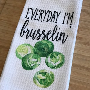 K&ouml;nnte beinhalten: Ein wei&szlig;es K&uuml;chentuch mit dem Text "EVERYDAY I'M brusselin'" in schwarzer Schrift. Das Handtuch zeigt eine Aquarell-Illustration von f&uuml;nf gr&uuml;nen Rosenkohl.