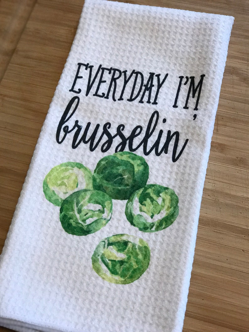 K&ouml;nnte beinhalten: Ein wei&szlig;es K&uuml;chentuch mit dem Text "EVERYDAY I'M brusselin'" in schwarzer Schrift. Das Handtuch zeigt eine Aquarell-Illustration von f&uuml;nf gr&uuml;nen Rosenkohl.