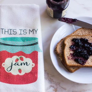 May include: A white kitchen towel with the text "This is my jam" and a red and blue illustration of a jar of jam. The towel is next to a plate with two slices of toast topped with blueberry jam.
