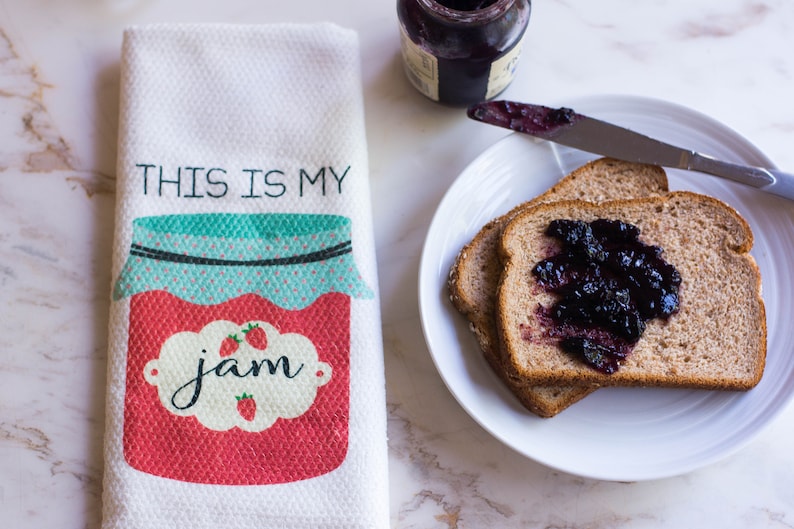 May include: A white kitchen towel with the text "This is my jam" and a red and blue illustration of a jar of jam. The towel is next to a plate with two slices of toast topped with blueberry jam.
