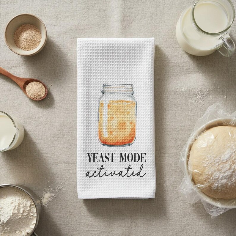 May include: White waffle-weave kitchen towel with a watercolor illustration of a jar filled with a golden liquid. The text "YEAST MODE activated" is printed below the jar in black script. Baking ingredients surround the towel.