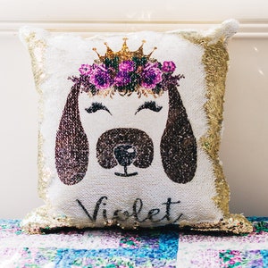 May include: A white and gold sequin cushion featuring a brown dog face with a purple flower crown. The dog's name, "Violet", is written beneath the face.
